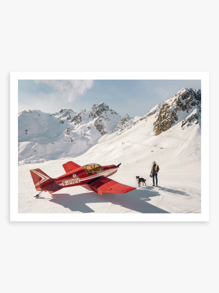 A fine art print by Paul Fuentes depicting a red airplane on a snowy mountain background with a person standing beside it.