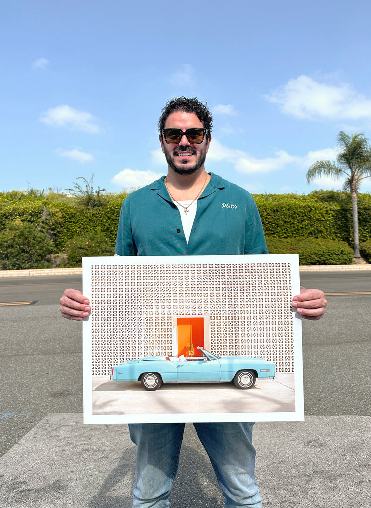Paul fuentes holding his fine art print of a light blue Cadillac parked in front of The Parker hotel in Palm Springs with the iconic breezy block wall.