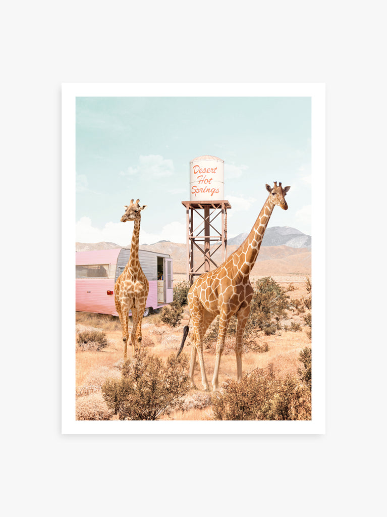 two giraffes in a desert setting with a water tower and a vintage-style pink trailer in the background. Fine art photography by Paul Fuentes
