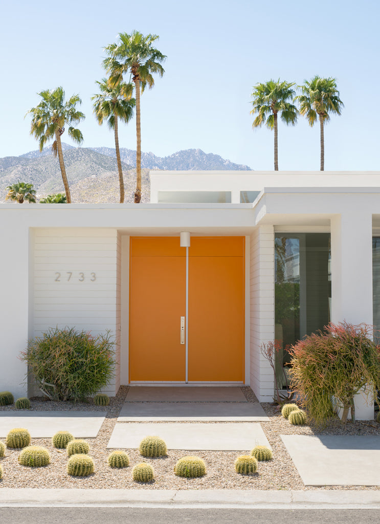 artwork featuring a vibrant orange front door set against a backdrop of palm trees and a white house with cactuses in palm springs, fine art photography by Paul Fuentes