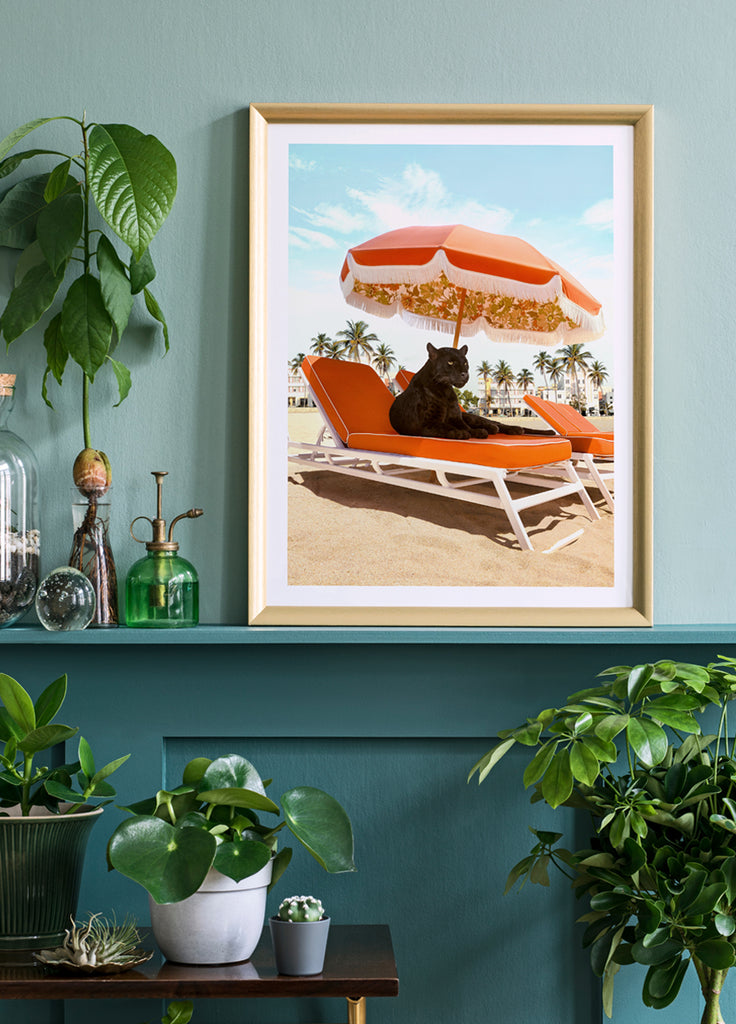 framed artwork of a retro beach vibe with a black panther lounging on an orange beach chair under a vintage  umbrella with palm trees in the background. fine art print by Paul Fuentes.