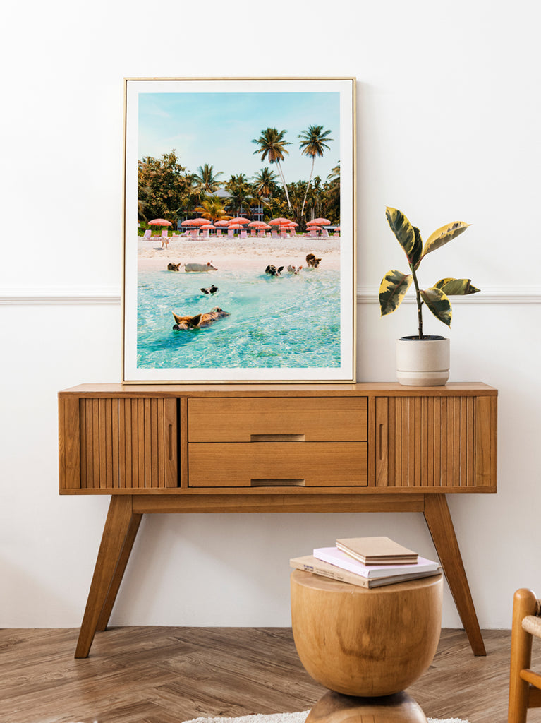 framed artworks by Paul Fuentes showing  pigs swimming in clear tropical water with palm trees and beach umbrellas in the background. on top of a wooden side table.